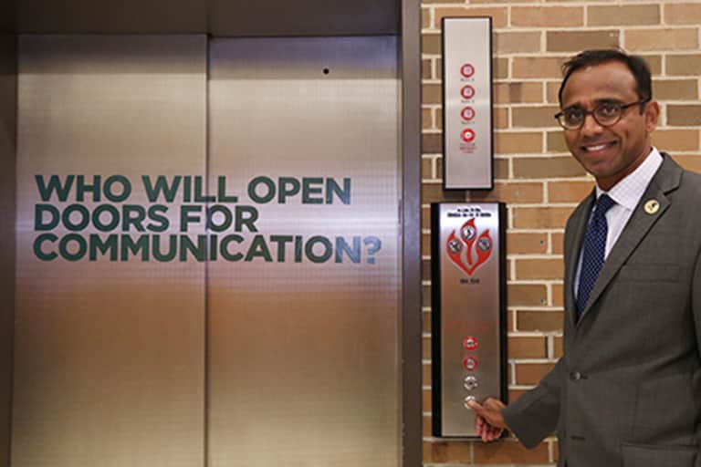 Dean Prabu David standing in front of the south lobby elevators. Elevator doors have sign on it that reads, who will open the doors for communication?