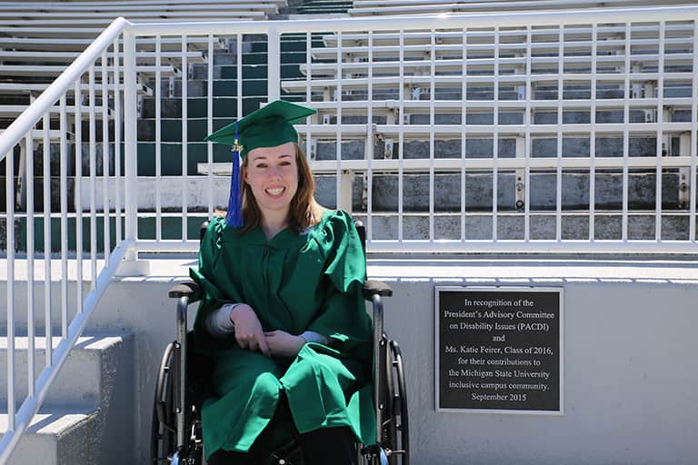Katie Feirer sitting in wheelchair at MSU Spartan Stadium next to the plaque. The plaque reads, "In recognition of the President's Advisory Committee on Disability Issues and Ms. Katie Feirer, Class of 2016, for their contributions to the MSU inclusive campus community. September 2015.