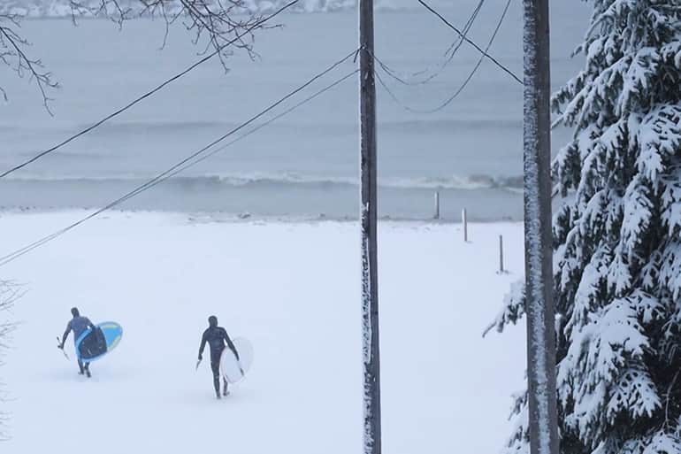 Two athletes walking across the snowy beachs of Michigan's Lake Huron in February preparing to paddle the freezing waves.