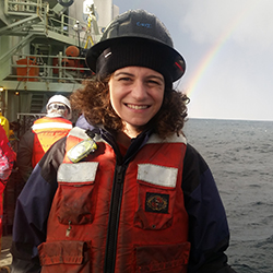 Stephanie Jordan on a ship wearing a life vest. A rainbow can be seen in the background behind her.