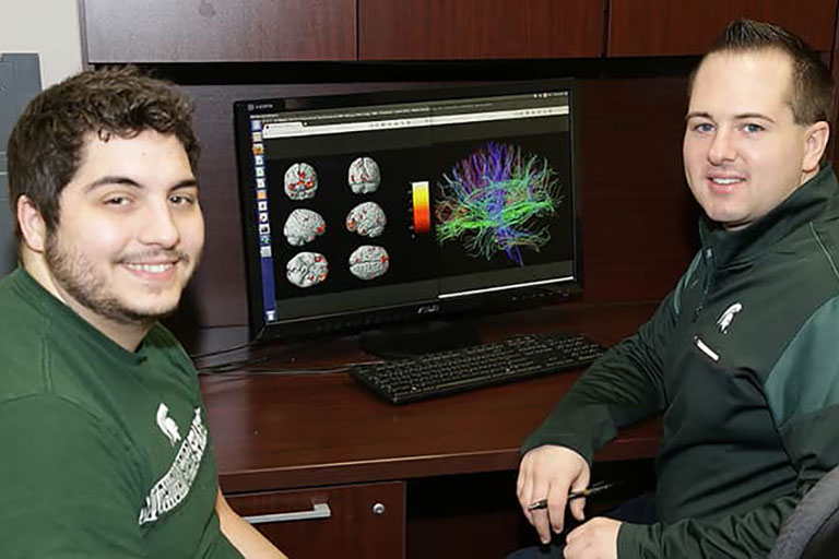 Two male CSD graduate students sitting down at a computer. Their computer monitor displays images of the brain from their recent research. 