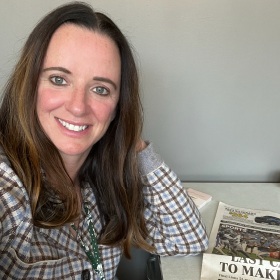 Tracy smiling wearing a plaid jacket with a newspaper on her desk. 