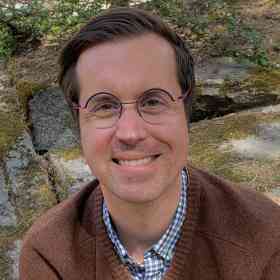 Close up of Ed Timke smiling and wearing circular glasses, a brown sweater. A mossy boulder is in the background.