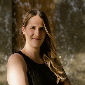 A woman with dark blonde hair in a black top stands in front of a waterfall.