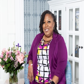 Black woman with a purple blazer, a black, white checkered and floral shirt with a gold necklace, sitting in a blue chair with flowers on the left hand side of her sitting on a table.