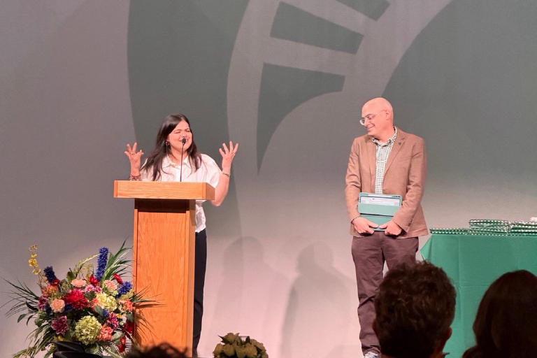 A person speaks at a wooden podium on a stage while another person stands nearby holding a folder. A floral arrangement sits beside the podium, and a table with awards is visible in the background. Audience members are seated in the foreground.