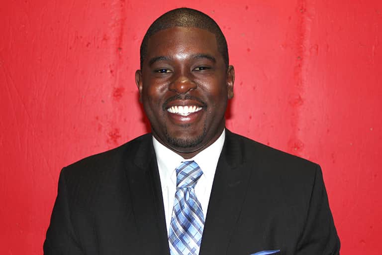 Jamal Spencer smiling dressed in dark suit and blue stripped tie against the background of red curtains.