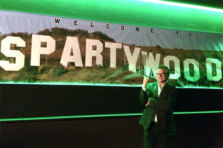 framed by vibrant green lights, a banner that looks like a photo of the "Hollywood" sign says "Spartywood" instead; Craig Murray points to it.